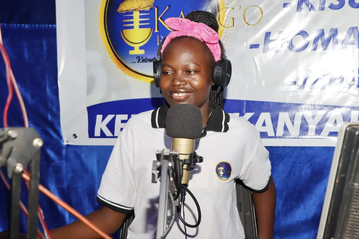 A presenter smiling at the studio microphone, headphones on, the Kang'o Radio banner behind her
