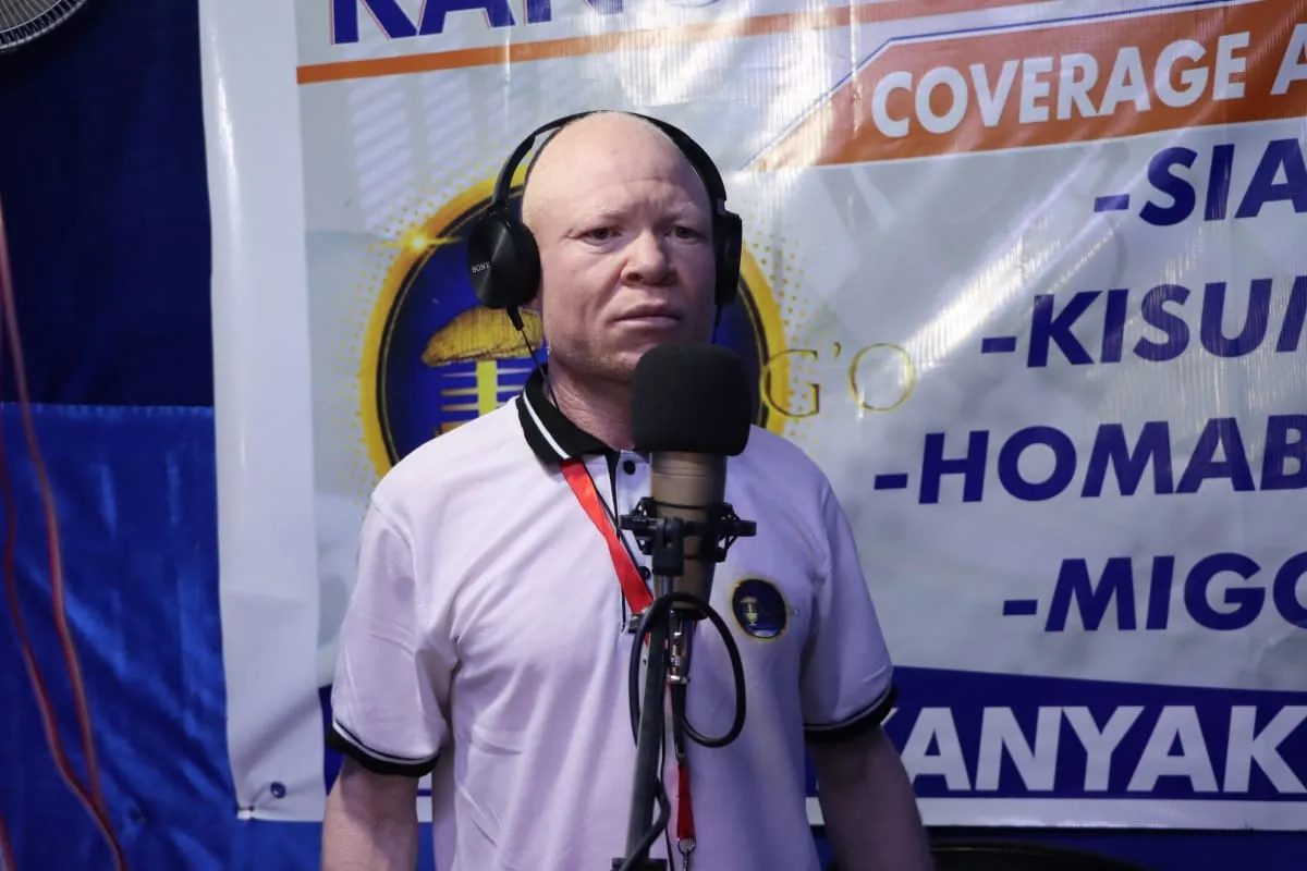 A presenter at the studio microphone, headphones on, in front of the station's banner